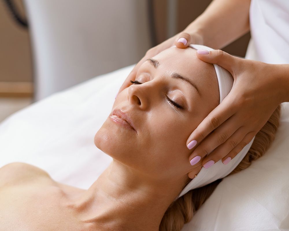 Una chica tumbada en la camilla recibiendo un masaje facial por una estilista.